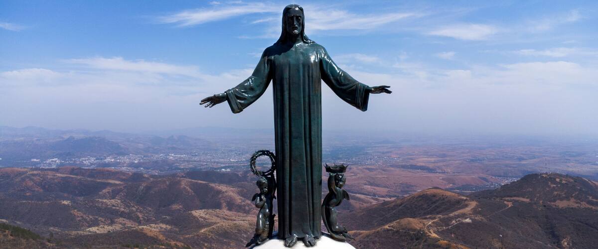 A statue of a robed figure with outstretched arms, flanked by two smaller figures, set against a mountainous backdrop.