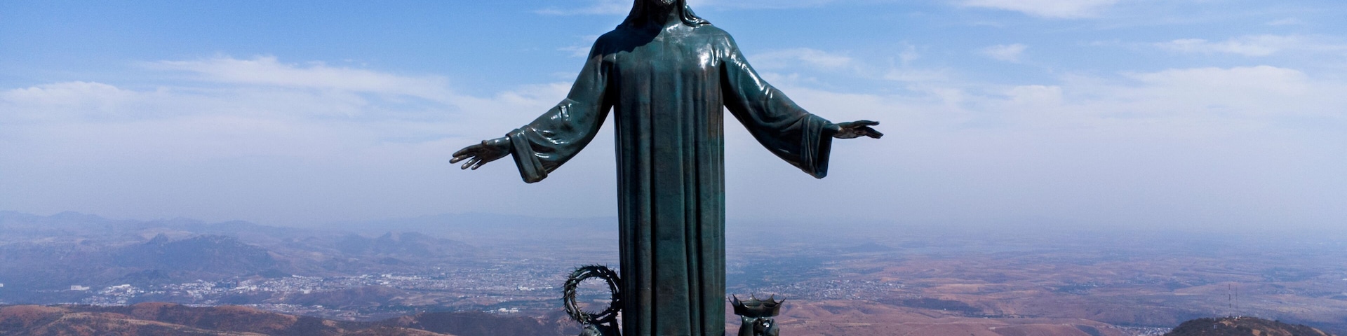 A statue of a robed figure with outstretched arms, flanked by two smaller figures, set against a mountainous backdrop.