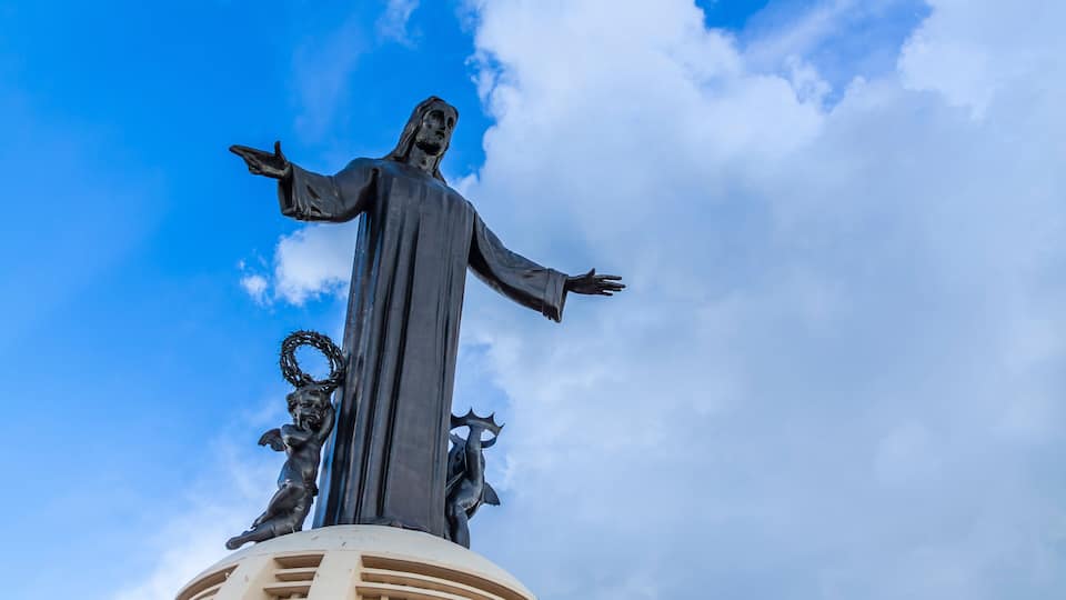 A statue of a robed figure with outstretched arms, flanked by two smaller figures, atop a rounded structure with a circular design, against a blue sky with clouds.