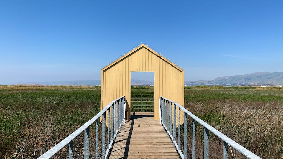 A wooden boardwalk leading to a yellow structure in a field with distant mountains.