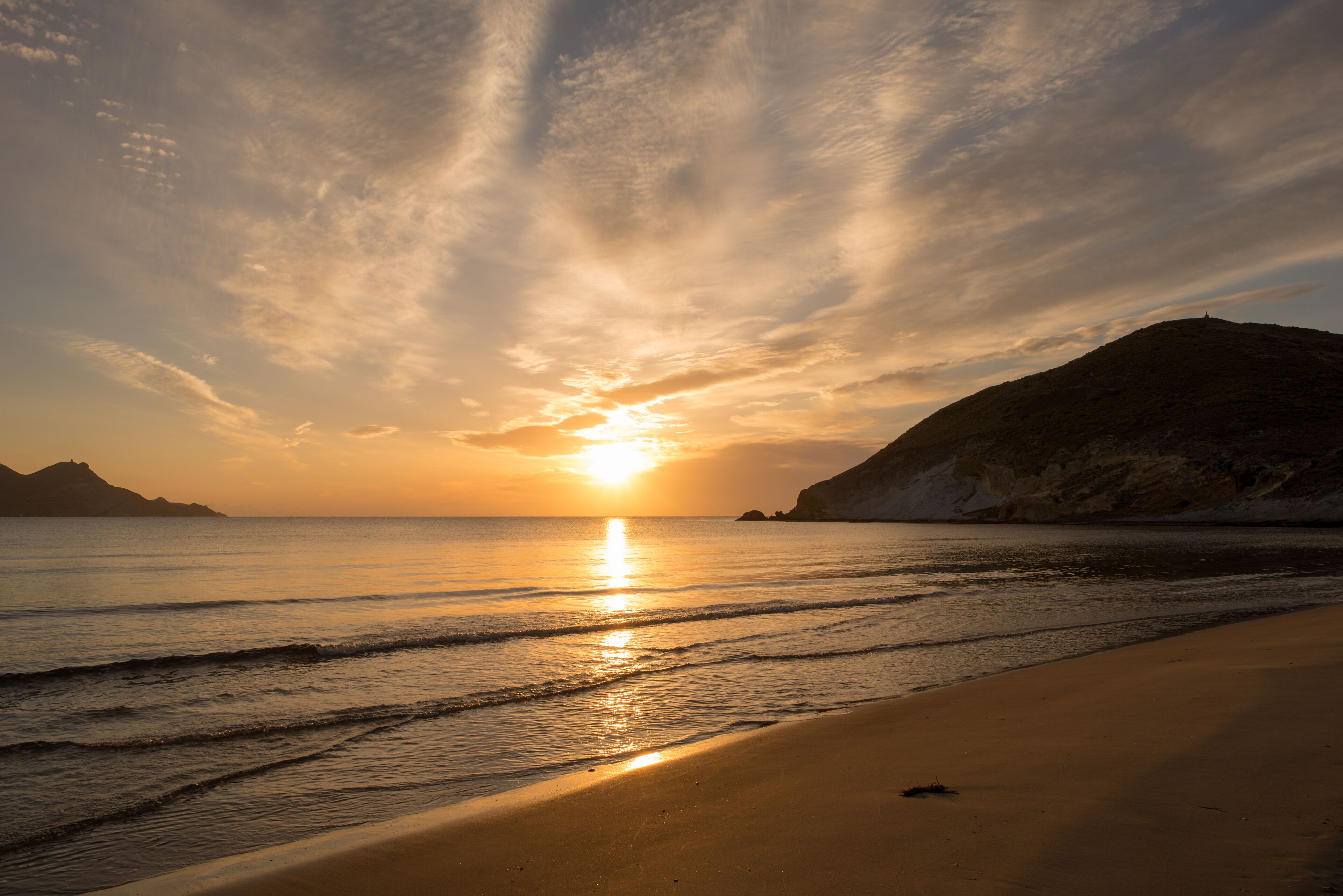Una puesta de sol sobre una playa con olas, una orilla arenosa y colinas a lo lejos.