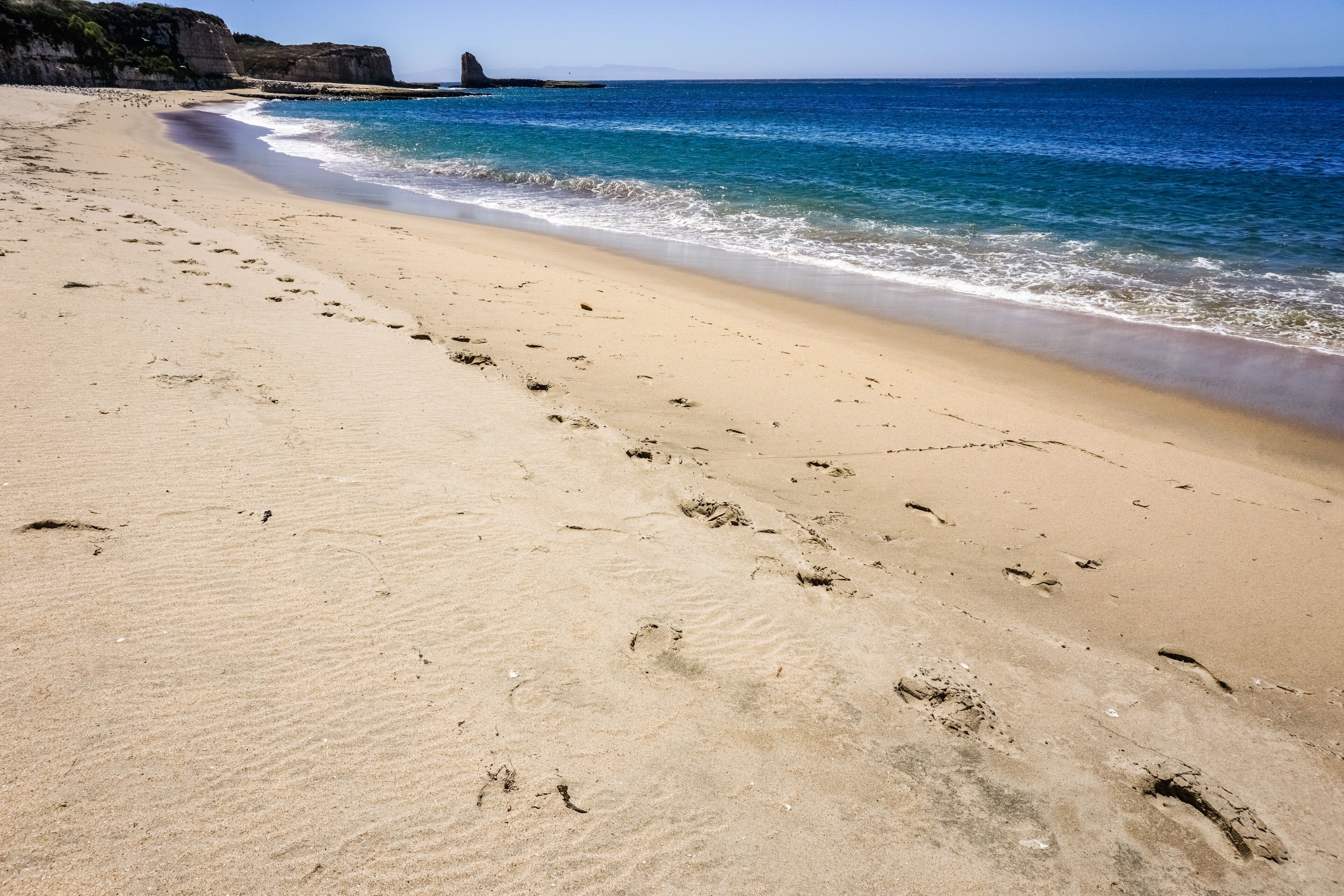 Una playa arenosa con huellas que conducen al agua, un acantilado rocoso al fondo y un cielo azul despejado.