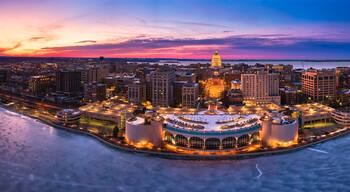 A cityscape at dusk with a prominent dome-shaped building, a waterfront, and a circular structure with multiple towers.