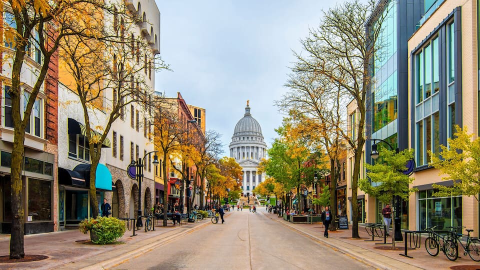 A city street lined with autumn-colored trees and historic buildings, leading towards a large domed capitol building.