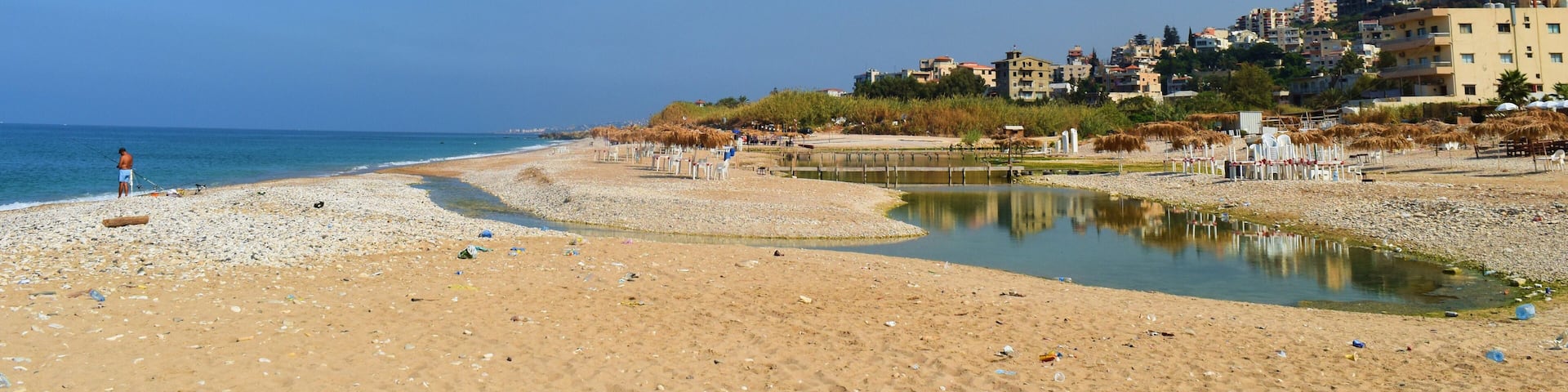 A beach with a small water body, pebbles, and buildings in the background.
