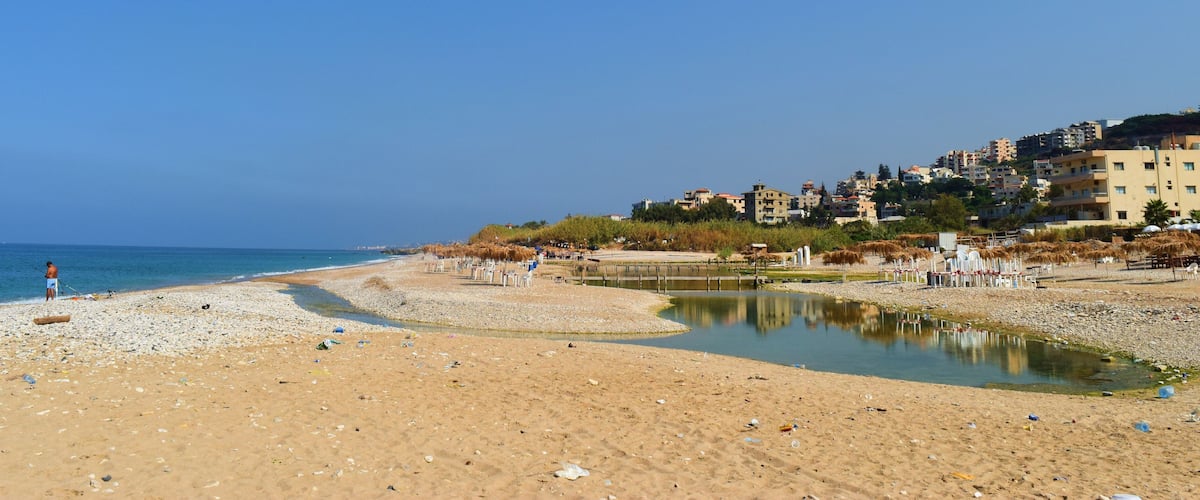A beach with a small water body, pebbles, and buildings in the background.