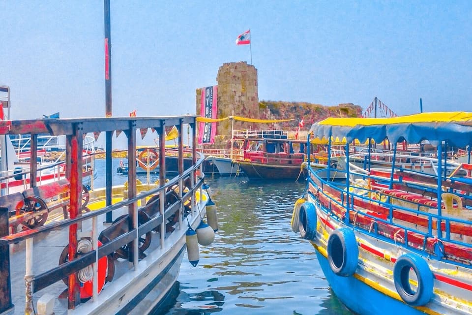 Un puerto con barcos de colores amarrados, un muelle con una bandera y una estructura que parece un castillo al fondo.