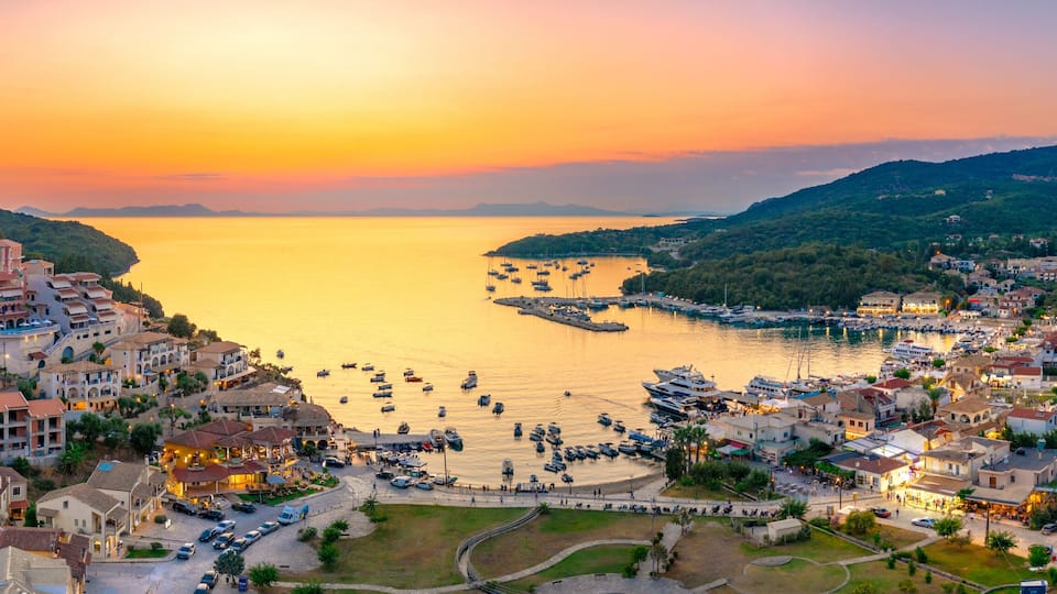 A coastal town at sunset with boats docked in the harbor and buildings along the hillside.