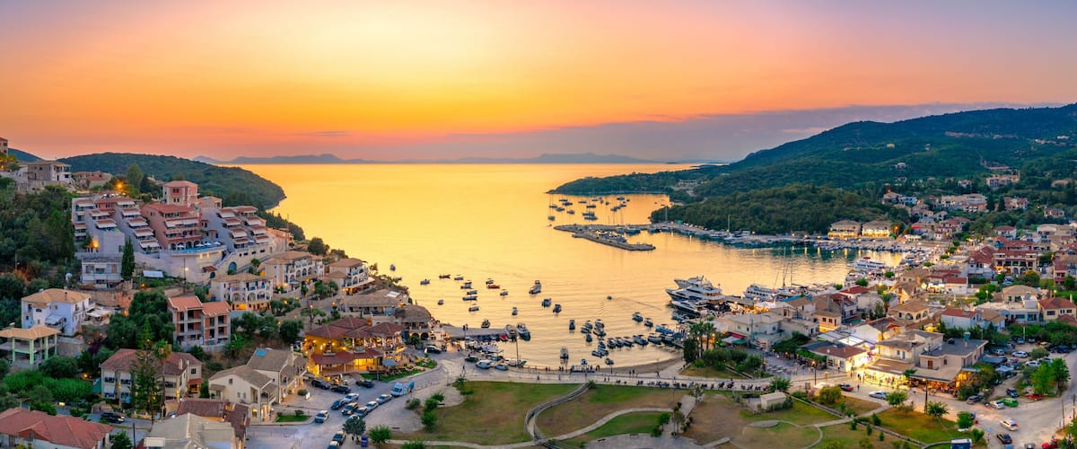A coastal town at sunset with boats docked in the harbor and buildings along the hillside.