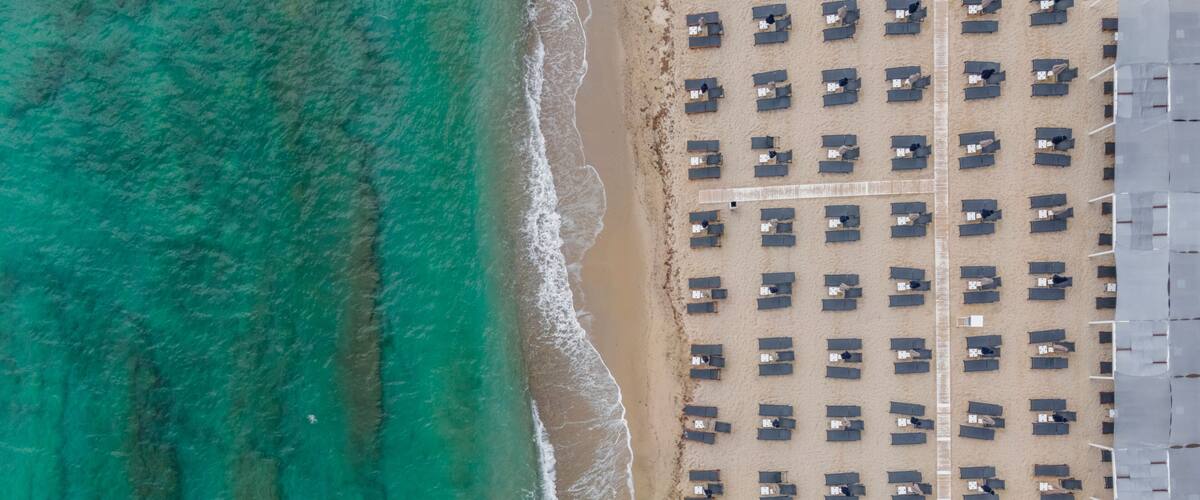 An aerial view of a beach with rows of beach huts and a clear turquoise sea.