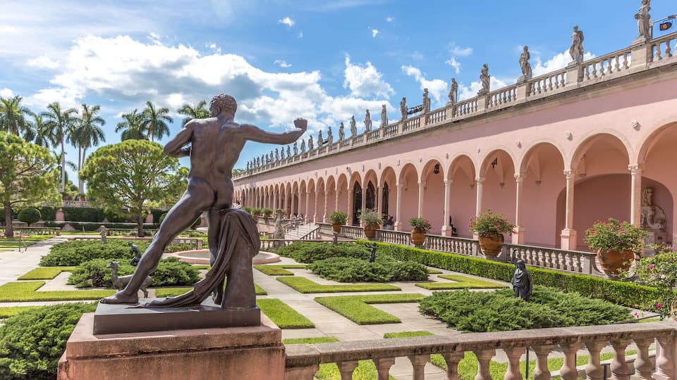 A statue of a man in a dynamic pose, a pink building with arches, and a well-maintained garden with hedges and palm trees.