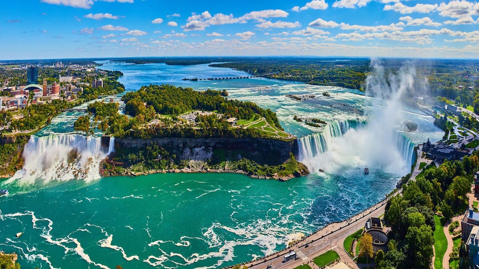 A large waterfall cascading into a turquoise river surrounded by greenery and urban areas.