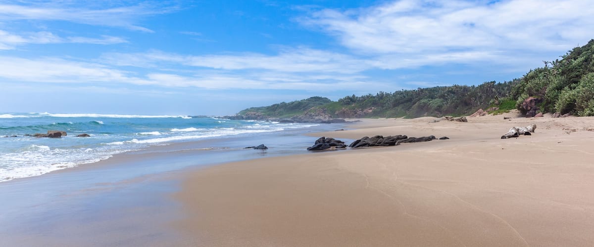 A sandy beach with waves, rocks, and a forested hillside.