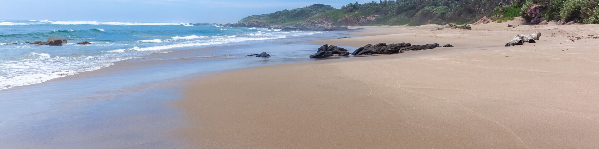 A sandy beach with waves, rocks, and a forested hillside.