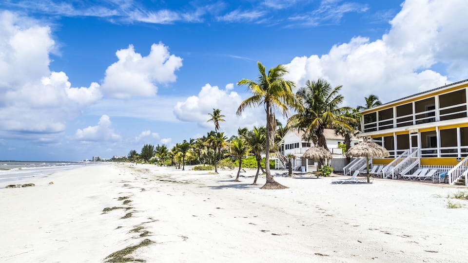 A beach with white sand, palm trees, and a yellow building with a staircase.