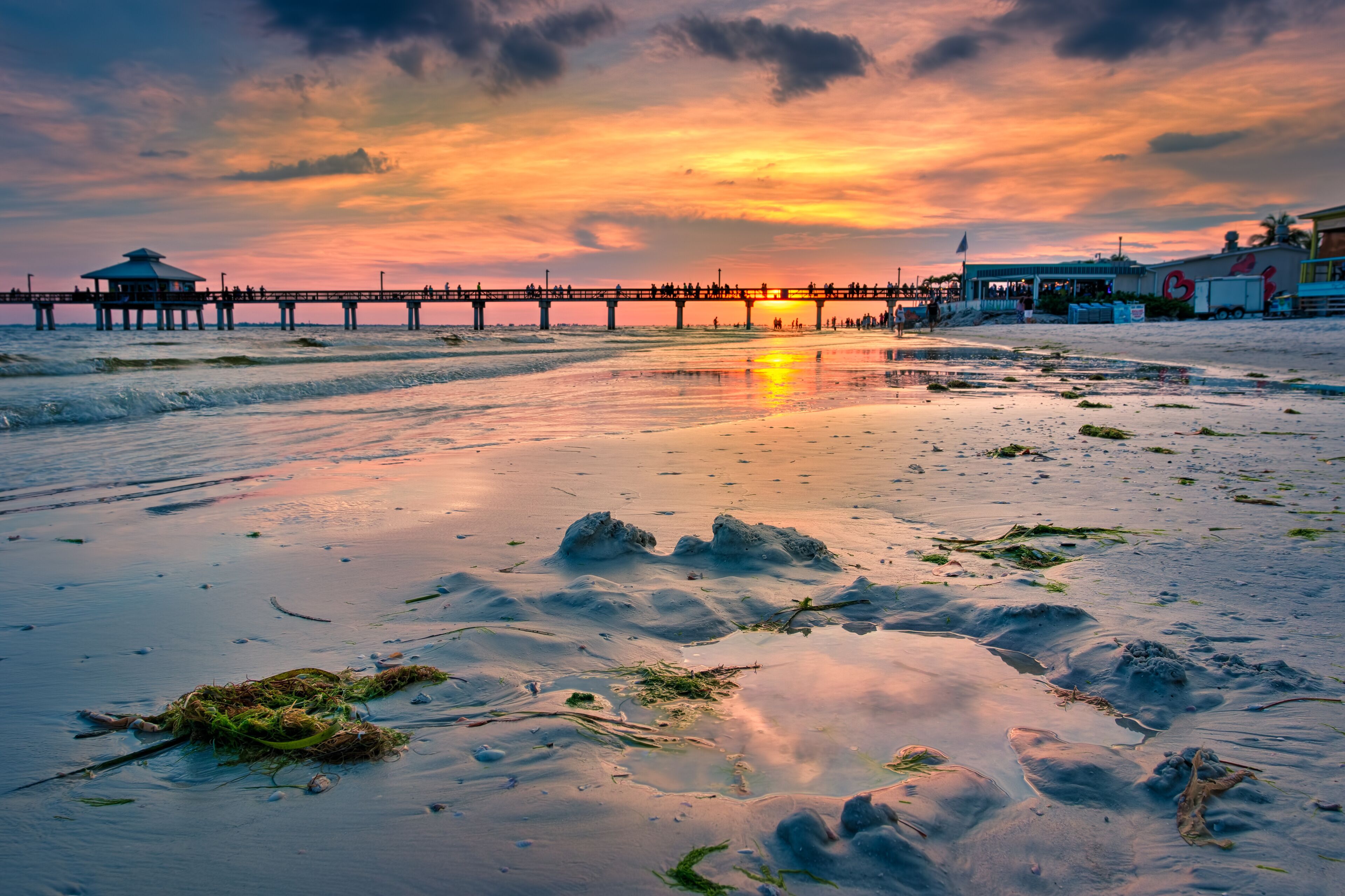 A sunset over a beach with a pier extending into the water, featuring a small structure on the left and a larger building on the right.