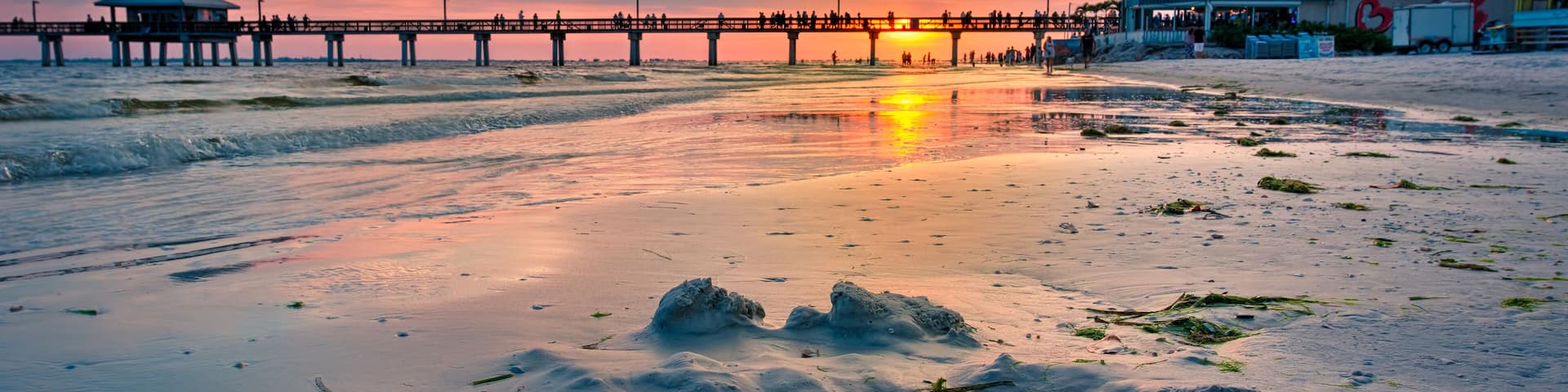 A sunset over a beach with a pier extending into the water, featuring a small structure on the left and a larger building on the right.