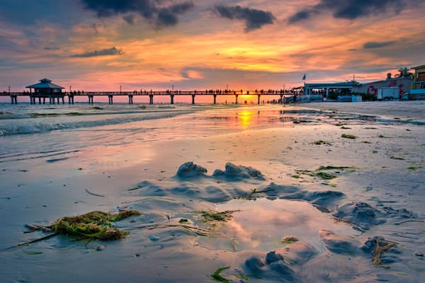 A sunset over a beach with a pier extending into the water, featuring a small structure on the left and a larger building on the right.