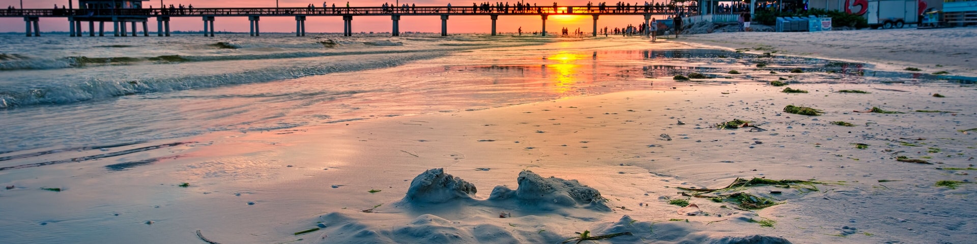 A sunset over a beach with a pier extending into the water, featuring a small structure on the left and a larger building on the right.