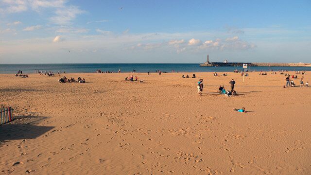 A wide sandy beach with people scattered around, a lighthouse in the distance, and a clear sky.