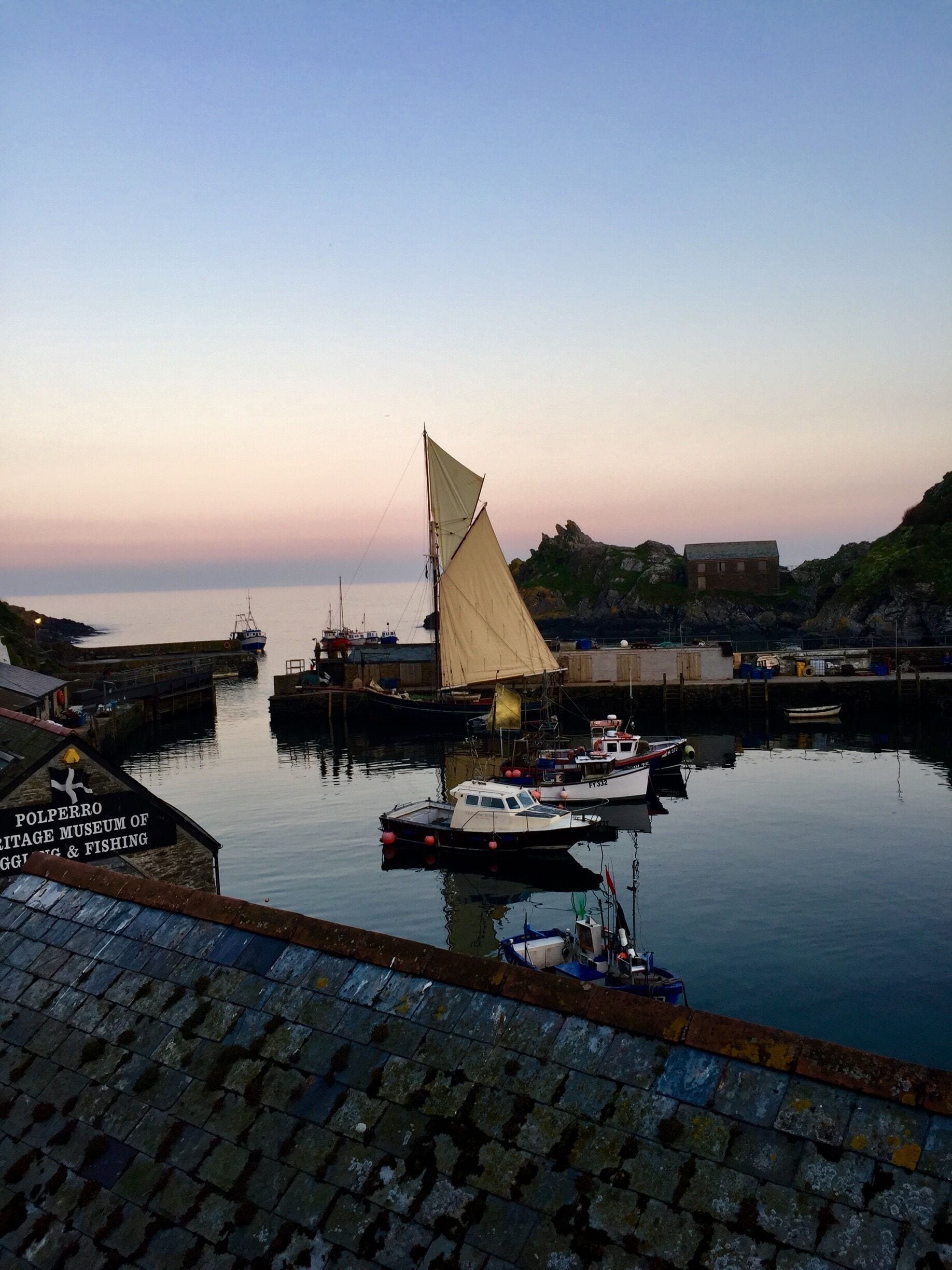 A sailboat with a large sail docked at a harbor with a sign for the Polperro Heritage Museum of Fishing & Fishing.