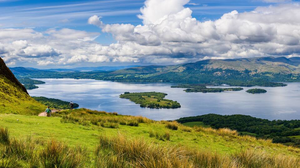 A lake surrounded by hills and a grassy area with two people walking.