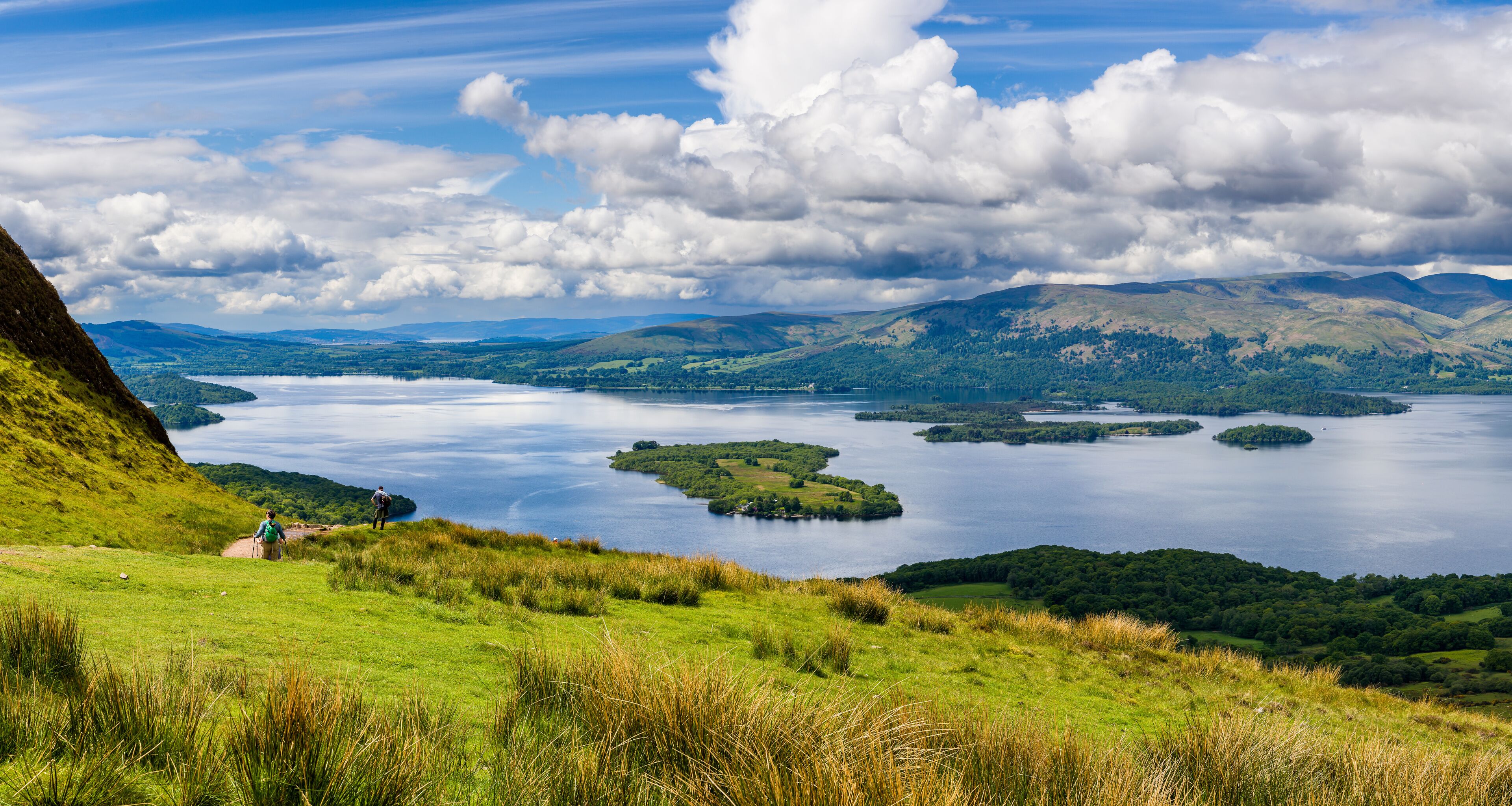 A lake surrounded by hills and a grassy area with two people walking.