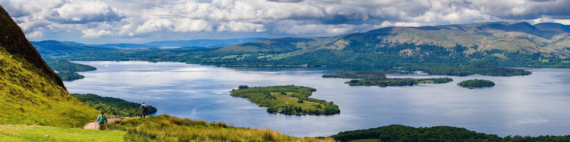 A lake surrounded by hills and a grassy area with two people walking.