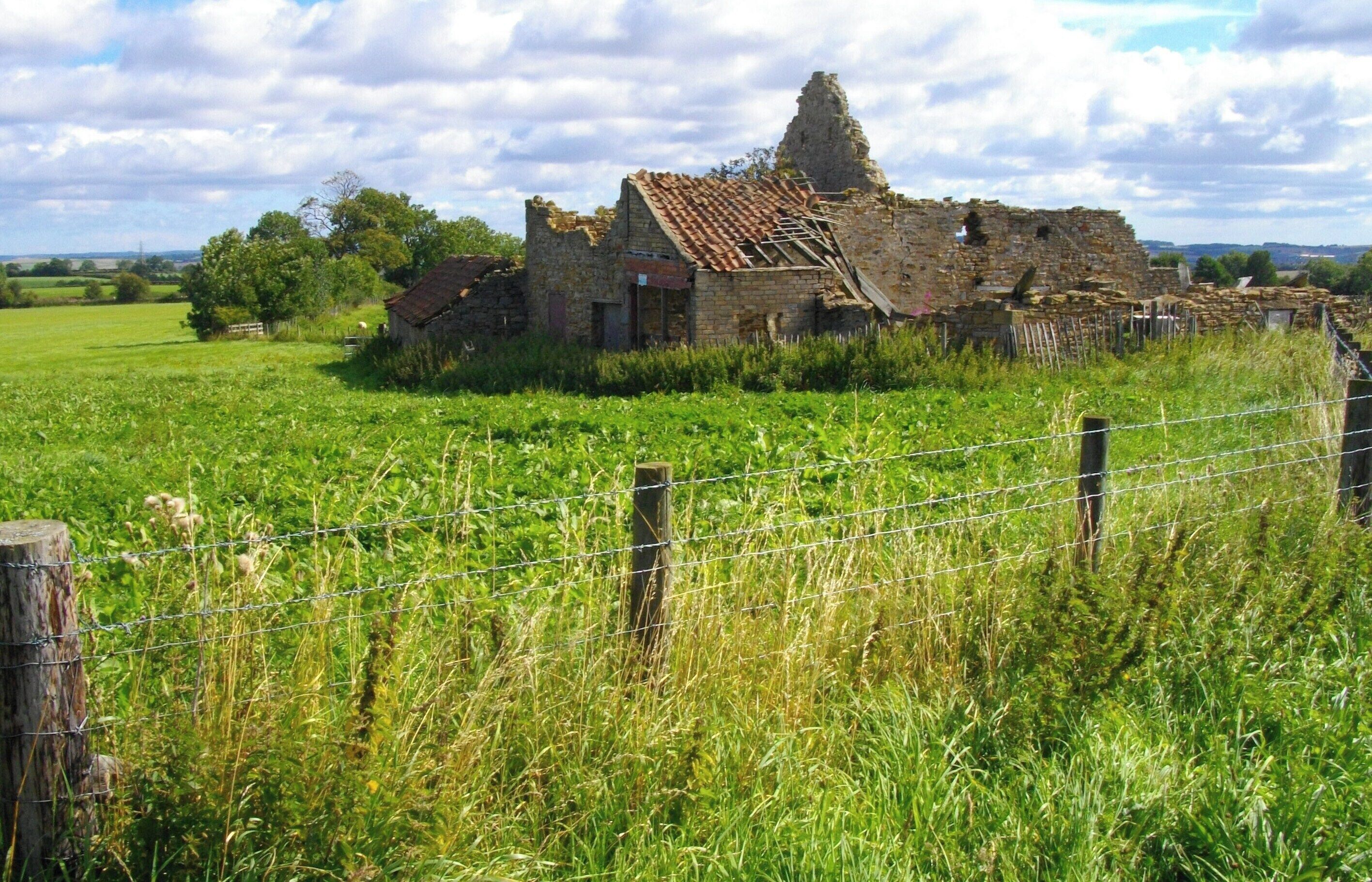 A dilapidated stone building with a tiled roof surrounded by a grassy field and a barbed wire fence.