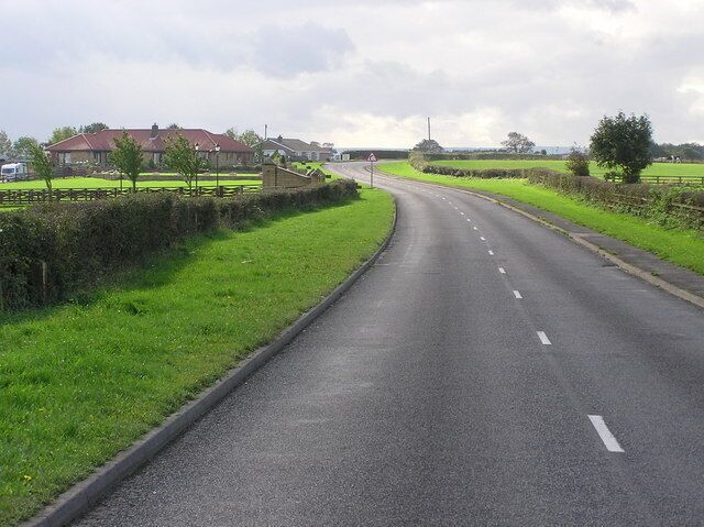 A rural road curving through green fields with houses in the distance.