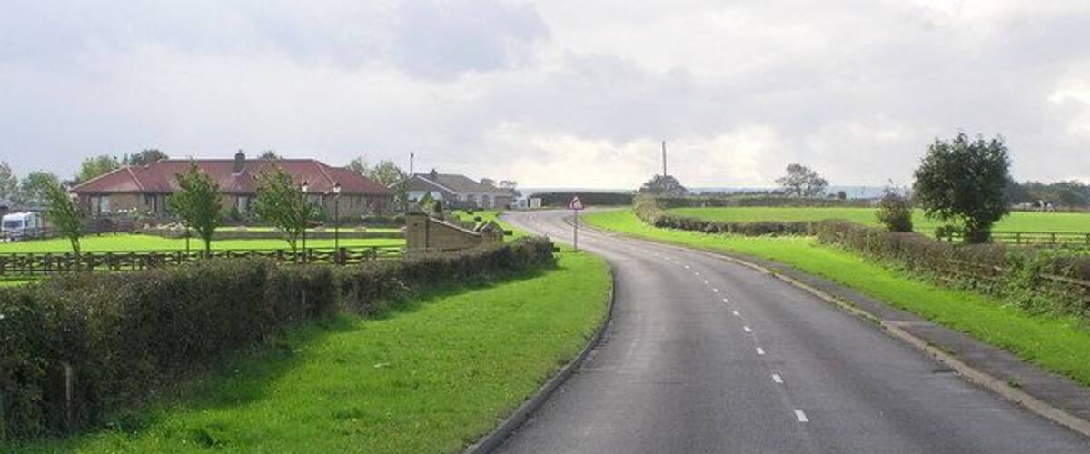A rural road curving through green fields with houses in the distance.