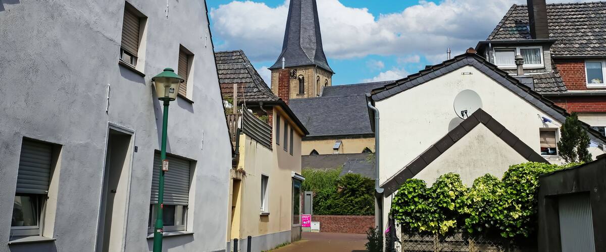 A narrow street with traditional European architecture, including a church with a tall steeple and a clock tower.