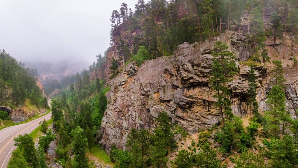 A winding road through a forested valley with a rocky cliff and a waterfall.