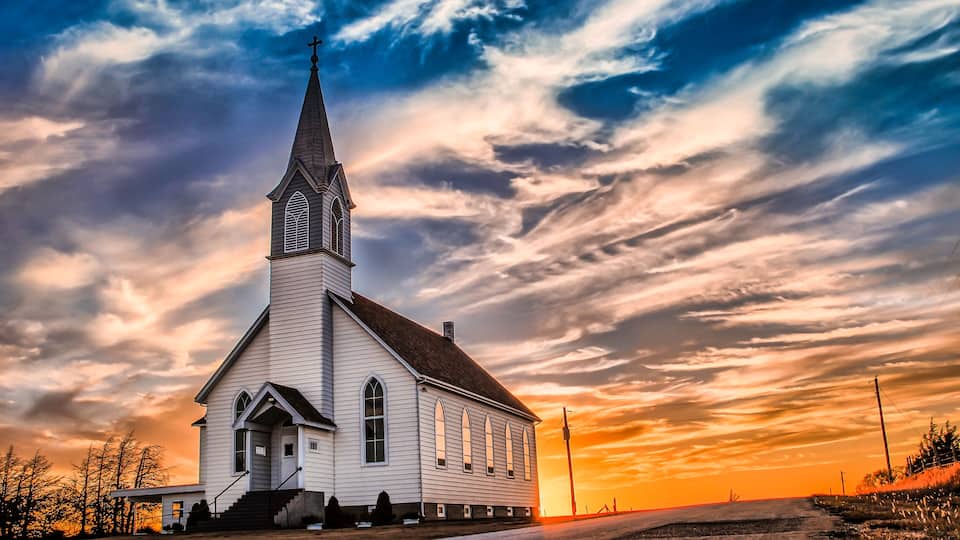 A white church with a tall steeple and arched windows, set against a dramatic sunset sky.
