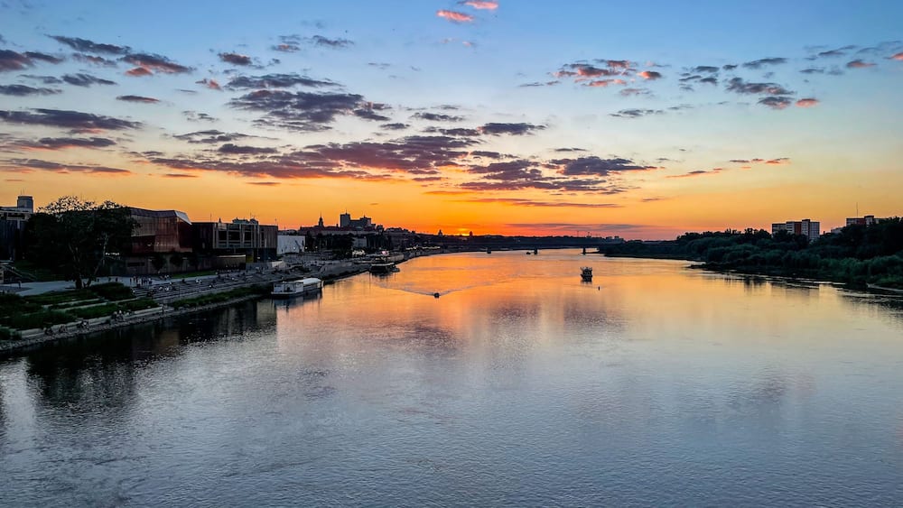 A river at sunset with a cityscape on the opposite bank, including modern buildings and a bridge.