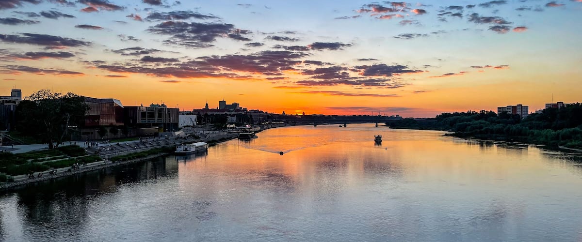 A river at sunset with a cityscape on the opposite bank, including modern buildings and a bridge.