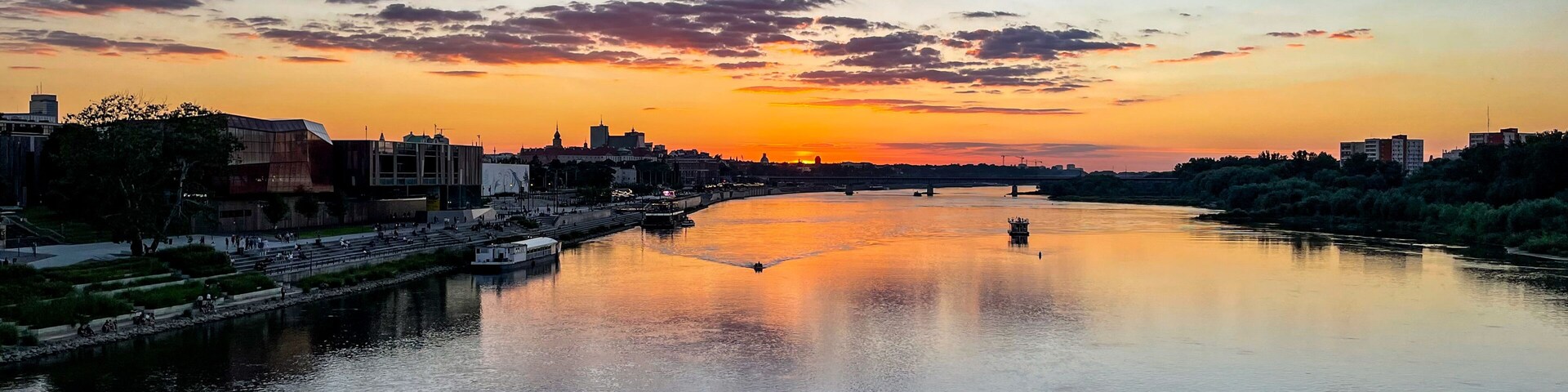 A river at sunset with a cityscape on the opposite bank, including modern buildings and a bridge.