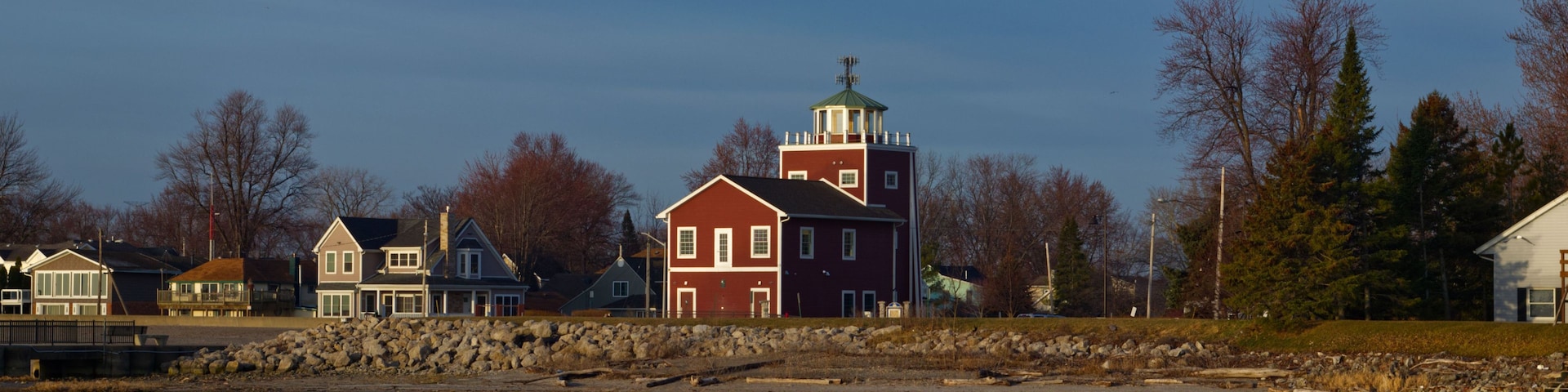 Un lago tranquilo que refleja un edificio rojo con torre, rodeado de árboles y otras casas.