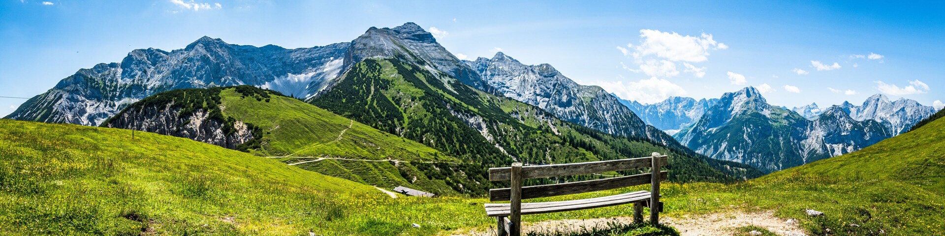 A wooden bench in a mountainous landscape with green hills and snow-capped peaks under a clear blue sky.