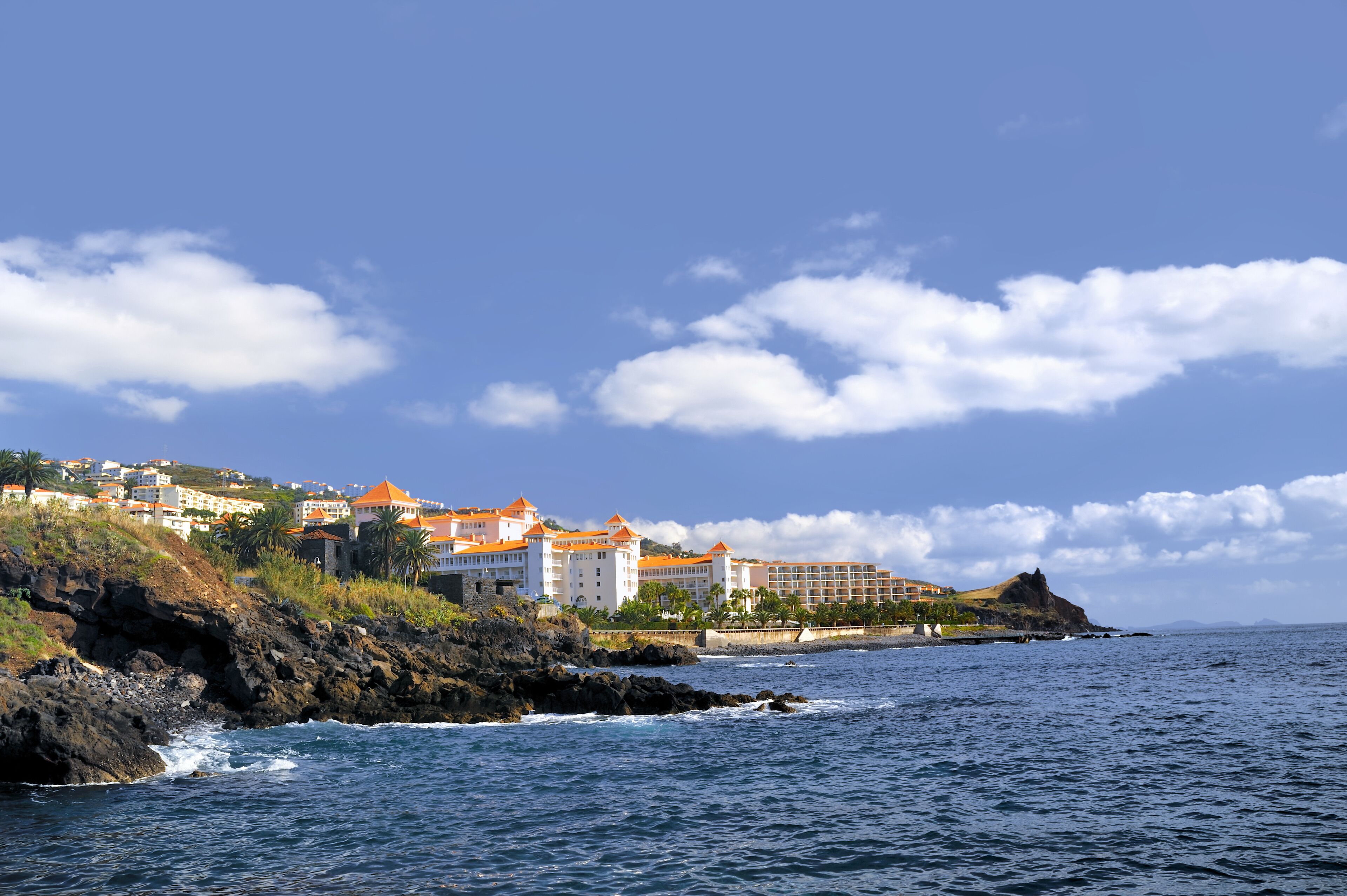 Um resort costeiro com edifícios numa encosta, costa rochosa e ondas do oceano.
