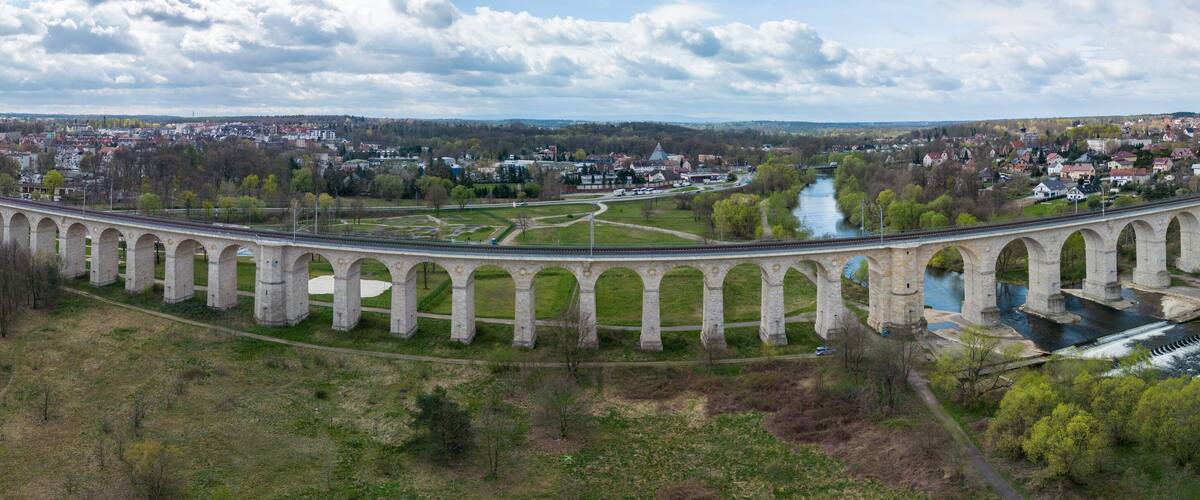 A stone viaduct spanning a river, surrounded by greenery and a town in the background.