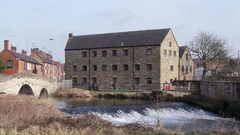 A stone building with a river flowing in front, a bridge, and residential buildings in the background.