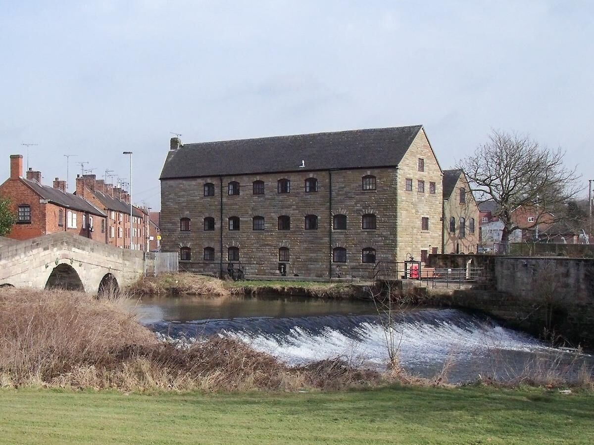 A stone building with a river flowing in front, a bridge, and residential buildings in the background.
