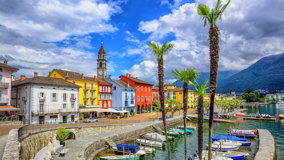 A colorful waterfront town with a prominent clock tower, boats docked along the canal, and lush palm trees.