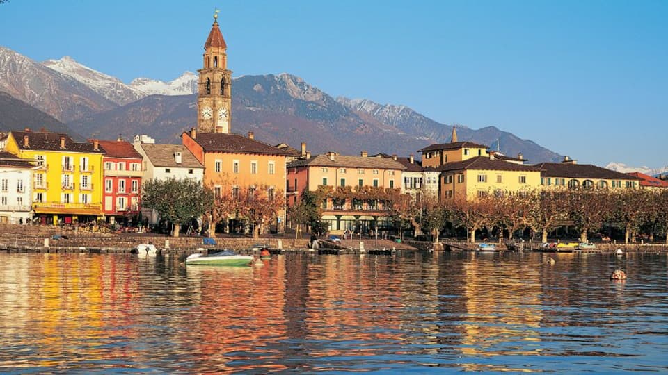 A lakeside town with colorful buildings, a prominent clock tower, and snow-capped mountains in the background.