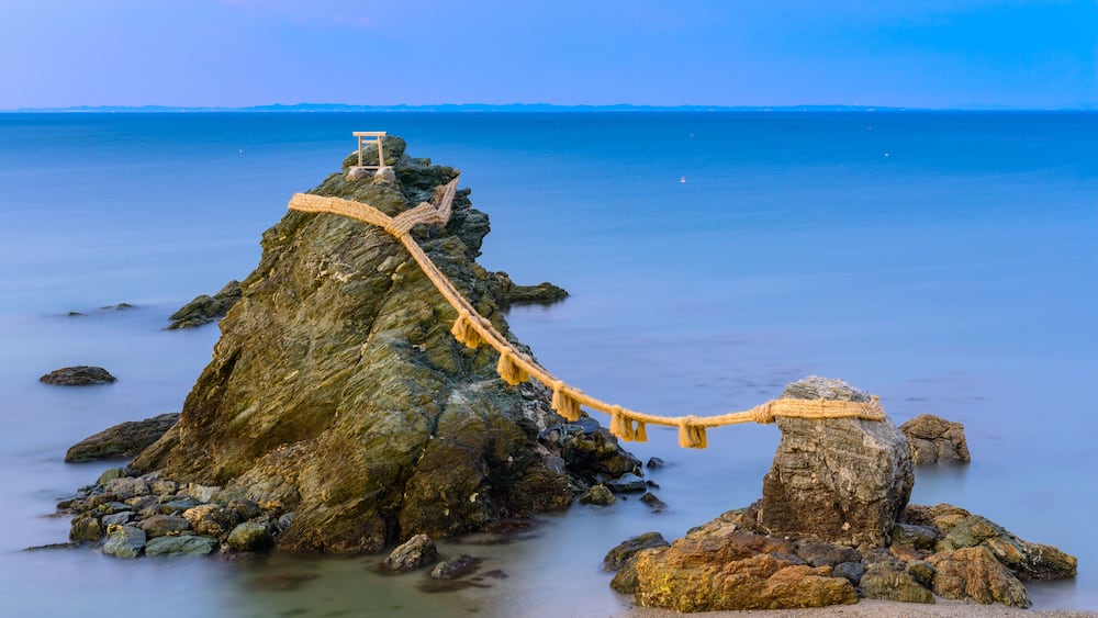 A rope bridge over a rocky island in a calm sea.