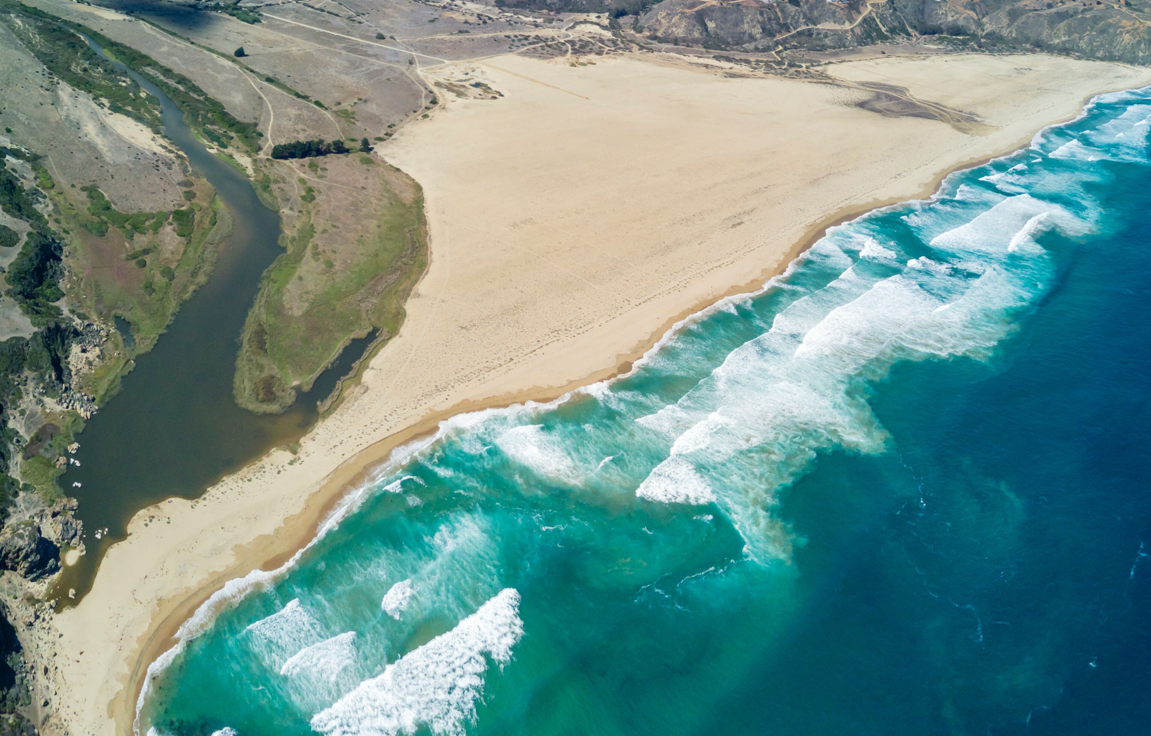 A coastal landscape with a sandy beach, a river flowing into the sea, and rocky terrain.