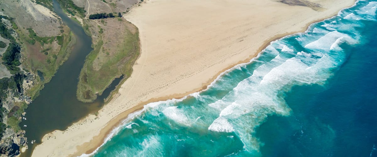 A coastal landscape with a sandy beach, a river flowing into the sea, and rocky terrain.