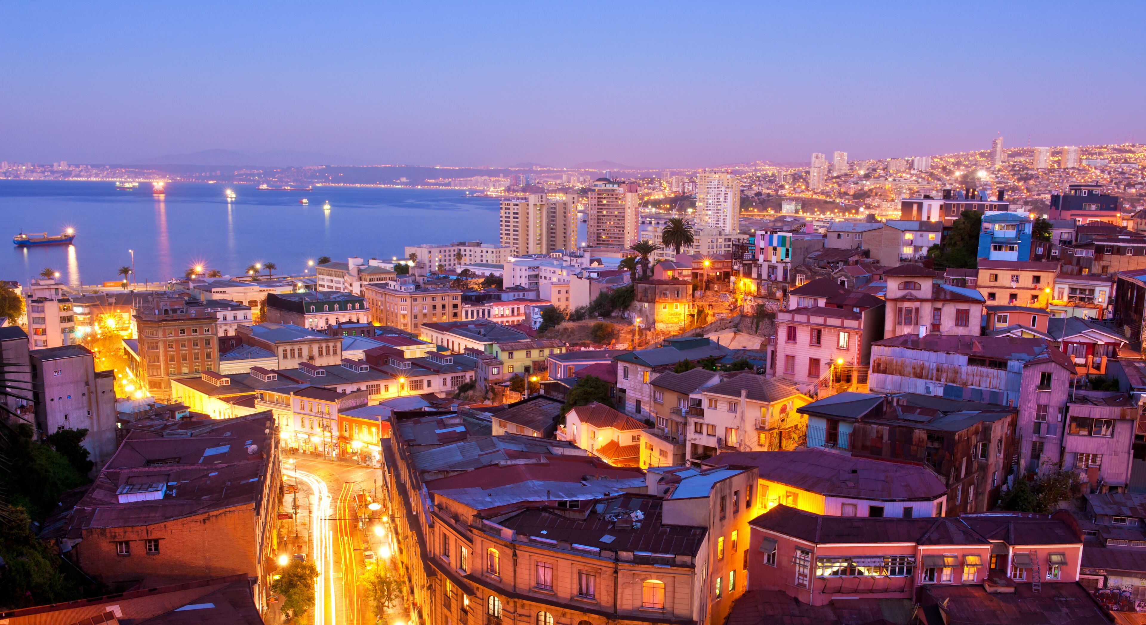 A cityscape at dusk with illuminated buildings and a river in the background.