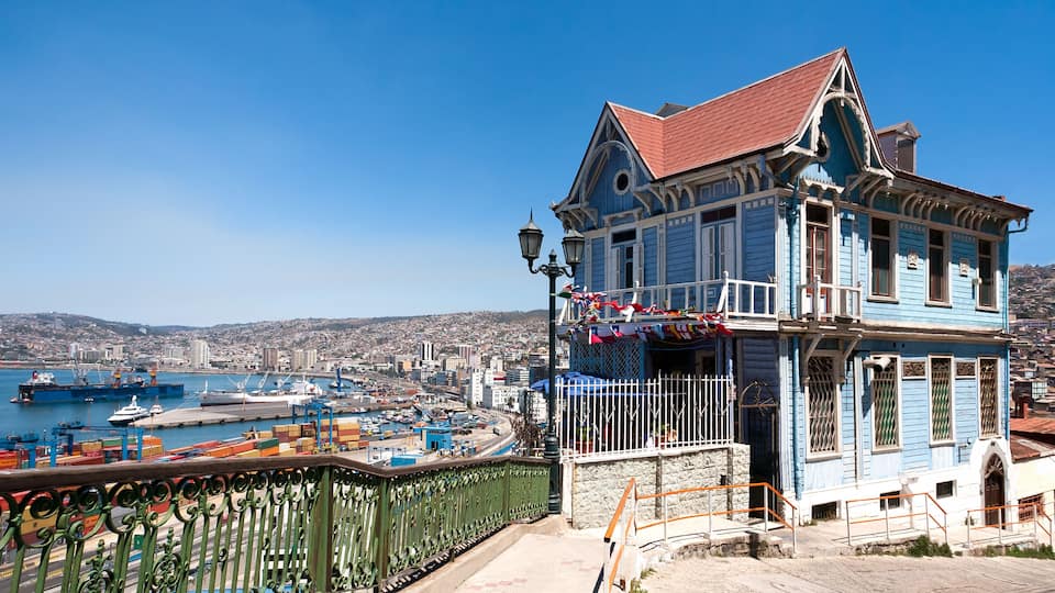 A blue house with a red roof overlooking a harbor with ships and a city in the background.